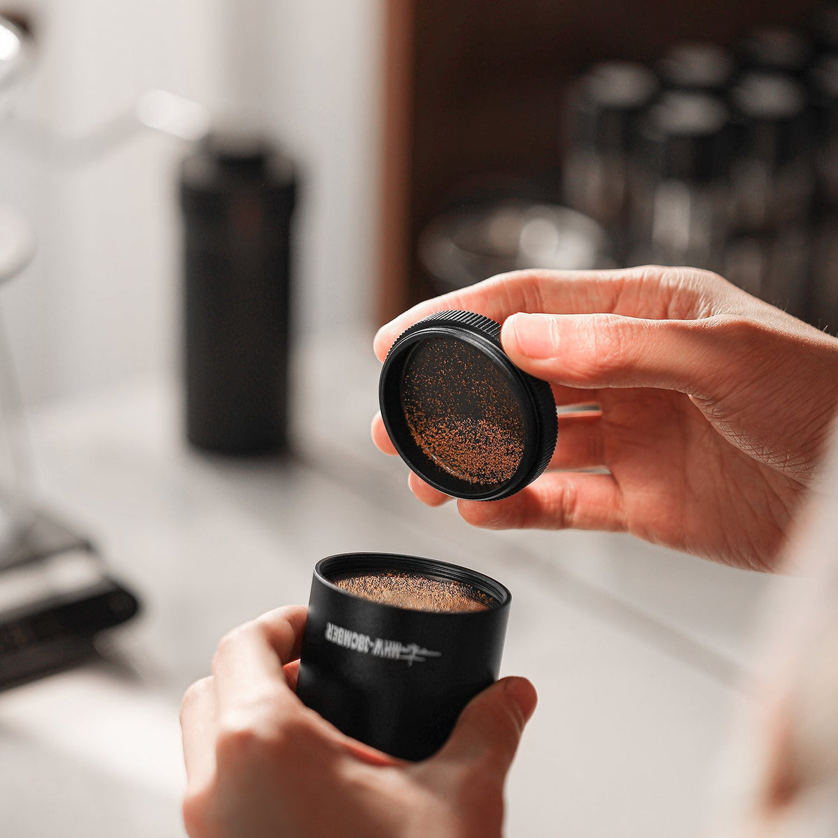 A person holds the MHW-3BOMBER Precision R3 Pro Manual Coffee Grinder (Black), unscrewing its lid to reveal finely ground coffee, with a blurred kitchen counter in the background.