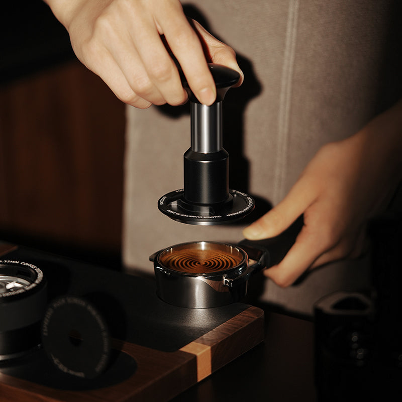 A person presses coffee grounds in a portafilter using the MHW-3BOMBER Knight Impact Tamper 58.35mm (Black), its 4-spring design ensuring even pressure under warm lighting, highlighting hands and textured grounds.