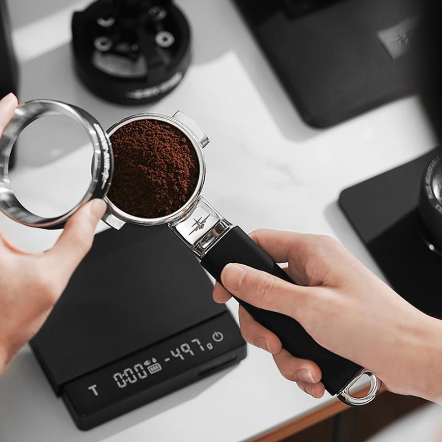 A person holds the MHW-3BOMBER YU Series Unibody Bottomless Portafilter 58mm over a digital scale showing 49.7 grams, preparing to tamp the grounds for optimal extraction.