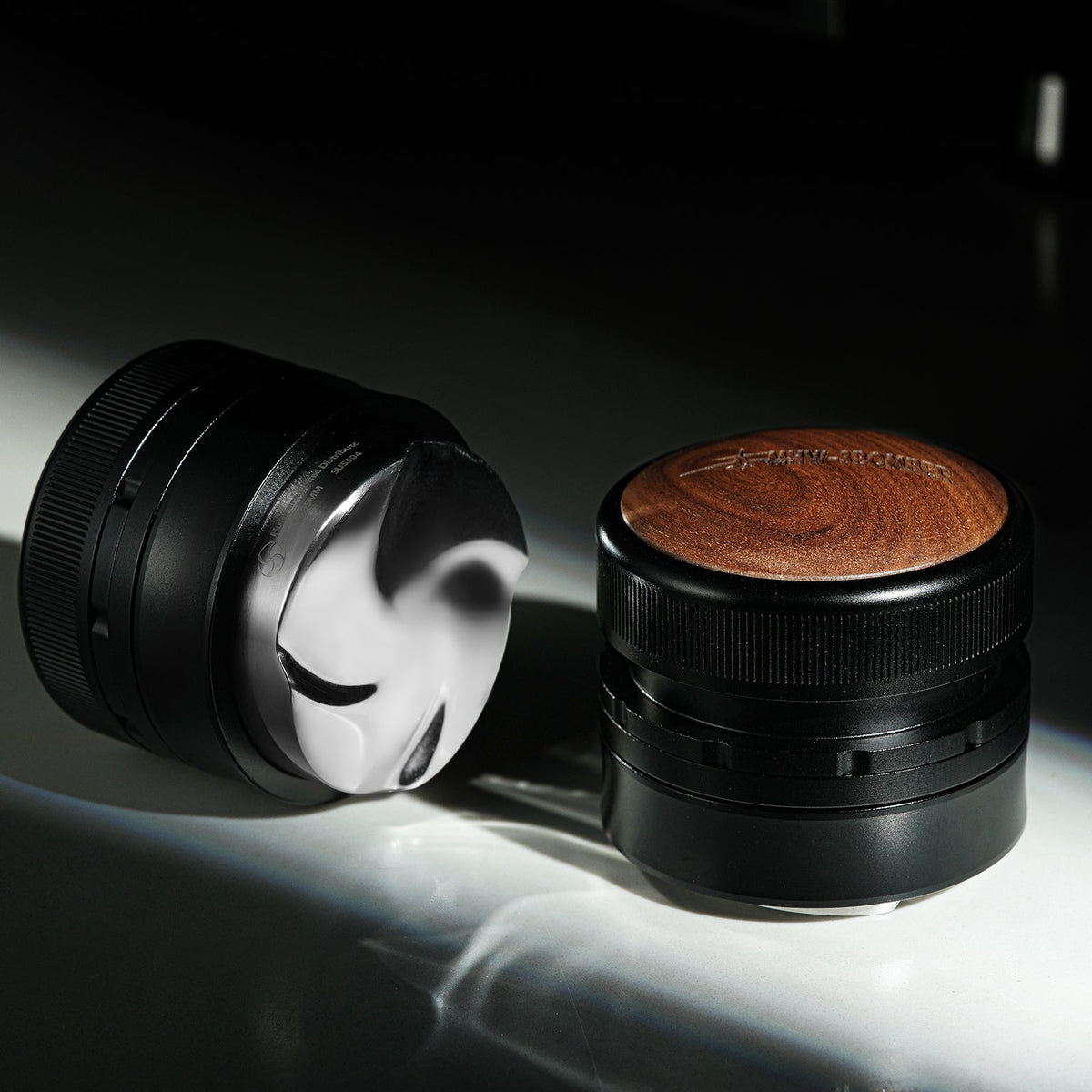 Two black cylindrical coffee tools, including an MHW-3BOMBER Coffee Gravity Distributor 58.35mm with a cyclone-shaped base, sit on a white surface with dramatic lighting and shadows.