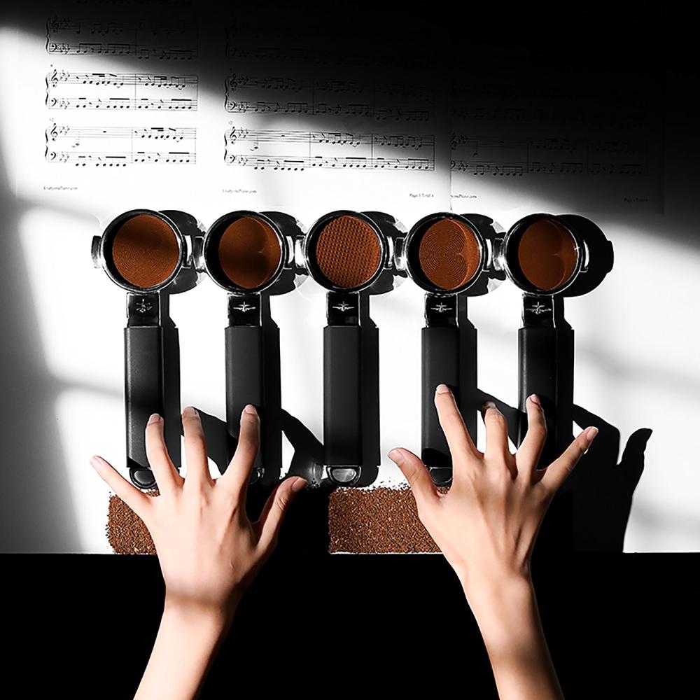 Two hands press coffee grounds with the MHW-3BOMBER YU Series Unibody Bottomless Portafilter 58mm, arranged like piano keys beneath sheet music, merging coffee and music while showcasing extraction efficiency.