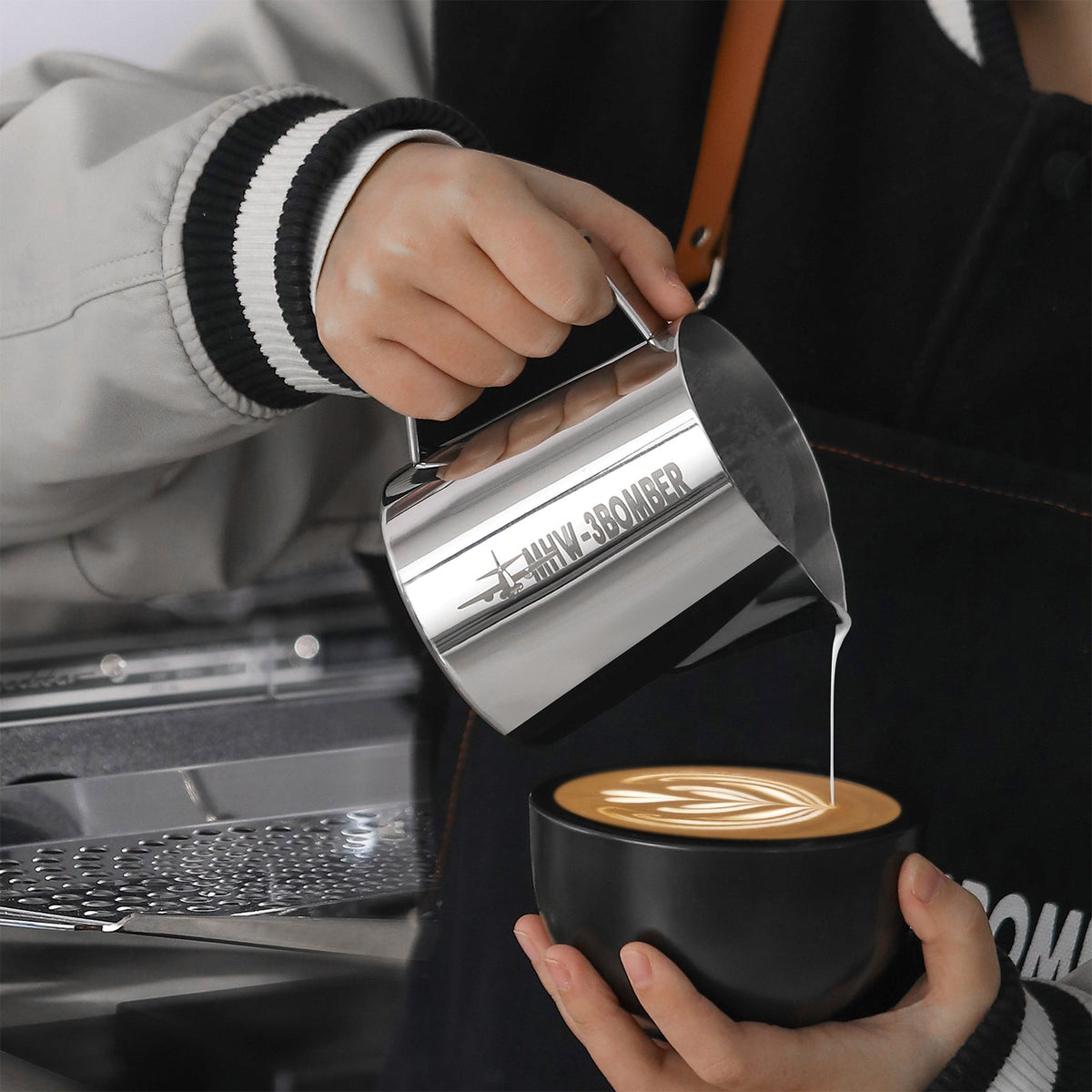 A person pours steamed milk from the MHW-3BOMBER Latte Art Milk Pitcher 5.0 500ml (Stainless Steel) into a black coffee cup, creating a latte art design on top.