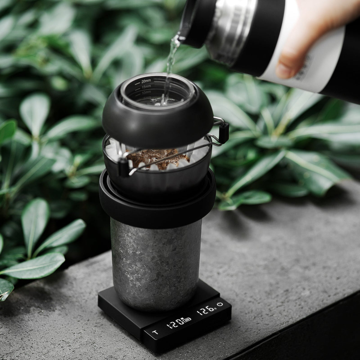 A hand pours hot water from a thermos into a coffee dripper set on a cup, which rests atop the MHW-3BOMBER Cube Coffee Scale 3.0 Micro, with green plants in the background.