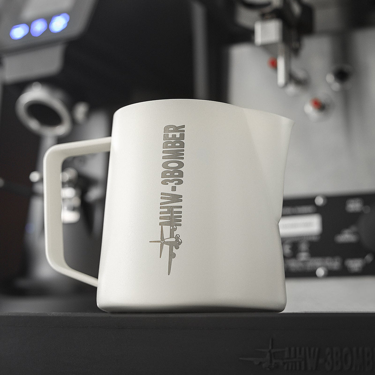 The MHW-3BOMBER Latte Art Milk Pitcher 5.0 500ml (White) sits on an espresso machine tray, featuring a plane graphic and “MHW-3BOMBER” branding, with blurred coffee equipment in the background.