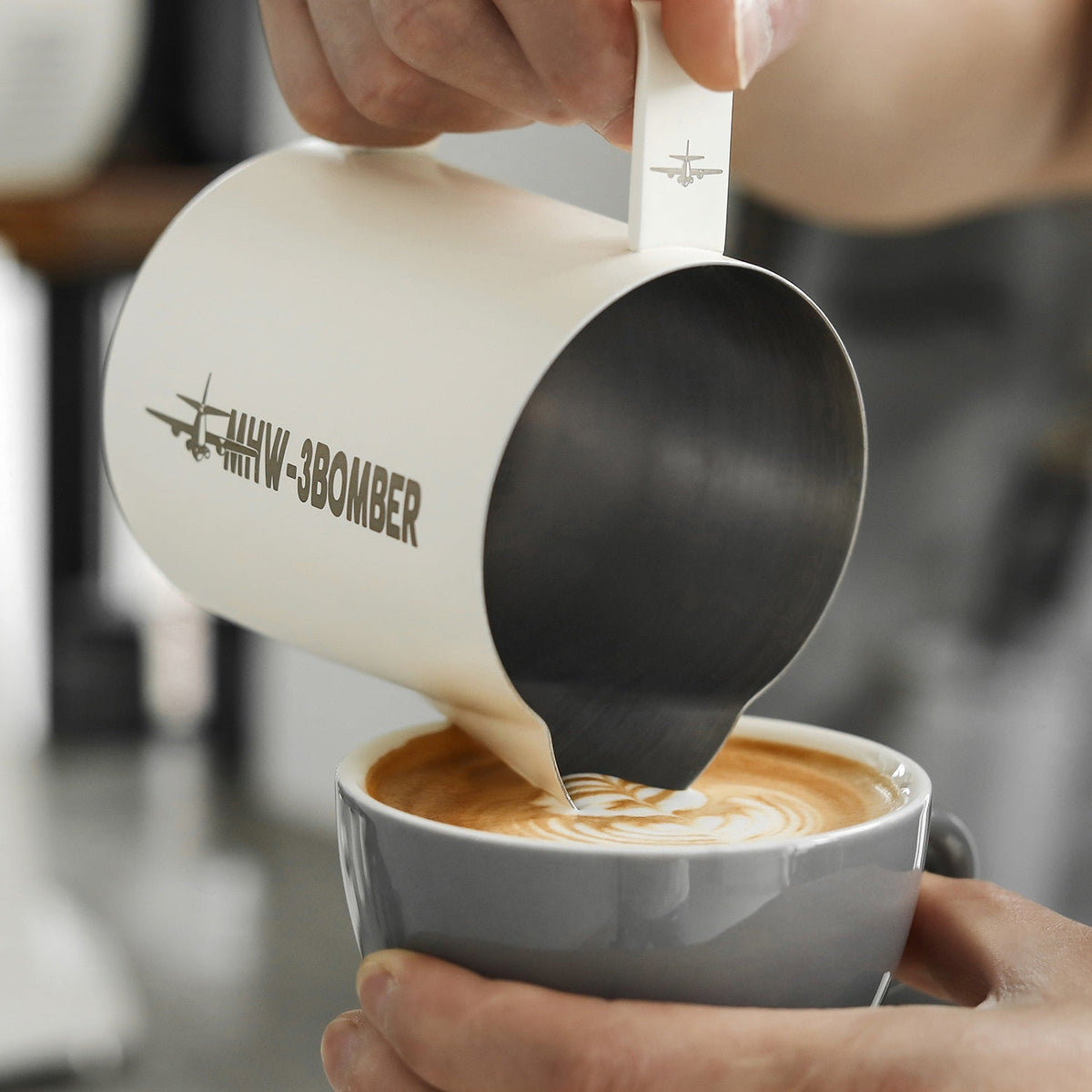 A hand pours steamed milk from an MHW-3BOMBER Latte Art Milk Pitcher 5.0 500ml (White) into coffee, using its patented spout to create latte art on the foam.