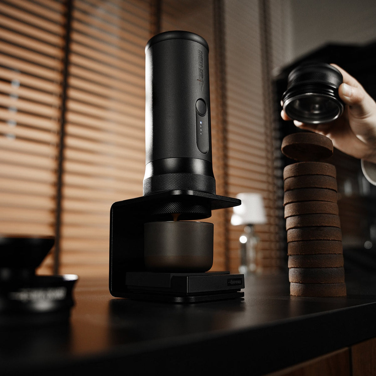 A sleek black coffee grinder sits by the MHW-3BOMBER X-press58 Portable Espresso Machine CM6954 (Black) and a neat stack of coffee pucks, while a hand holds a lid above them. Blinds and soft lighting are in the background.