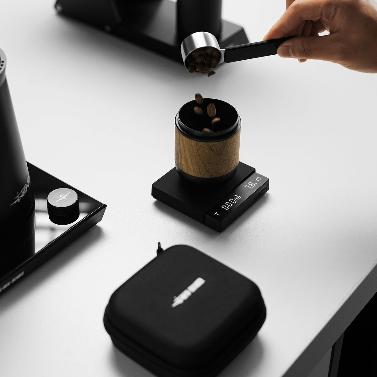 A hand pours coffee beans from a scoop into a small container positioned on the MHW-3BOMBER Cube Coffee Scale 3.0 Micro. The precision digital scale by MHW-3BOMBER displays numbers, while a black zippered case sits nearby on a white surface.