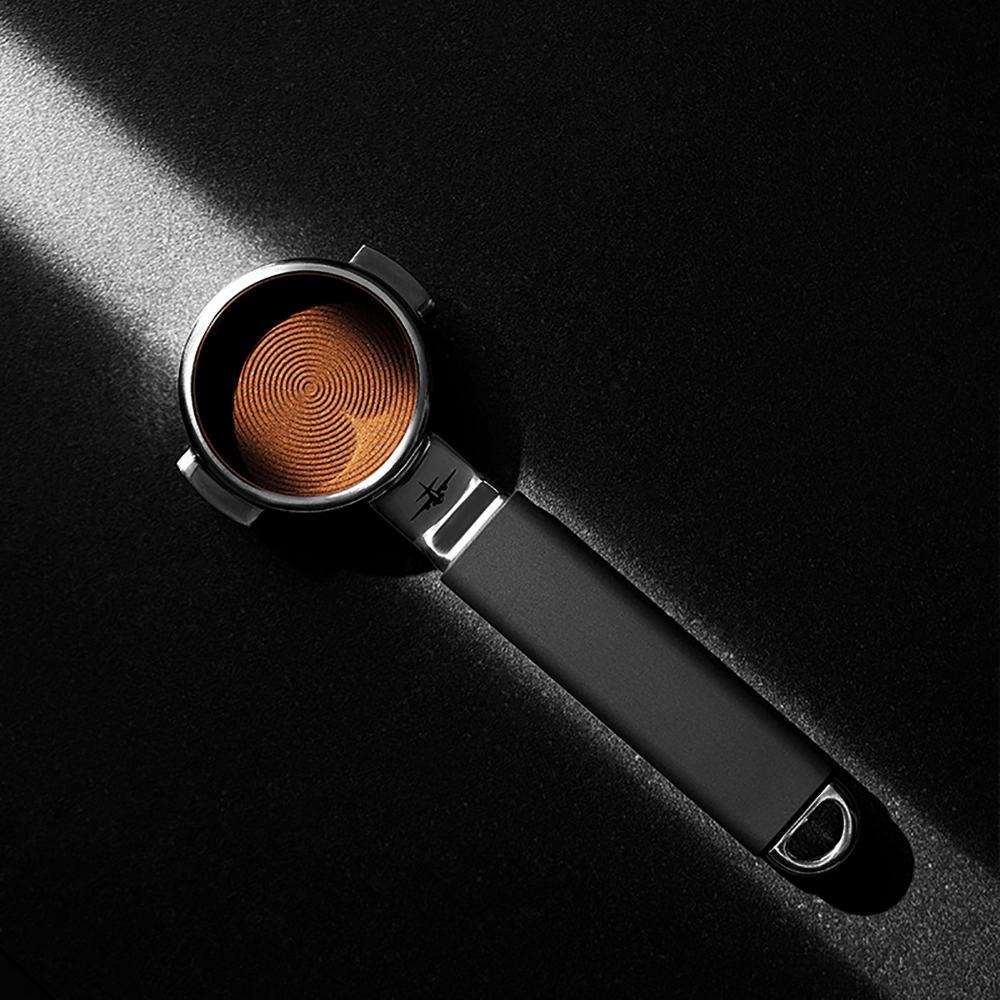 The MHW-3BOMBER YU Series Unibody Bottomless Portafilter 58mm, filled with espresso grounds and showing a spiral pattern, rests on a dark surface with a beam of light illuminating part of it.