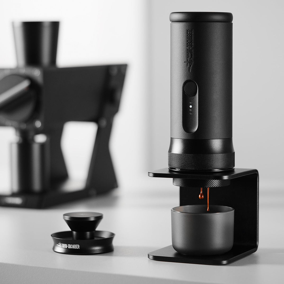 The MHW-3BOMBER X-press58 Portable Espresso Machine CM6954 (Black) dispenses coffee into a cup on its stand, with another coffee device and accessories blurred in a modern, minimalistic background.