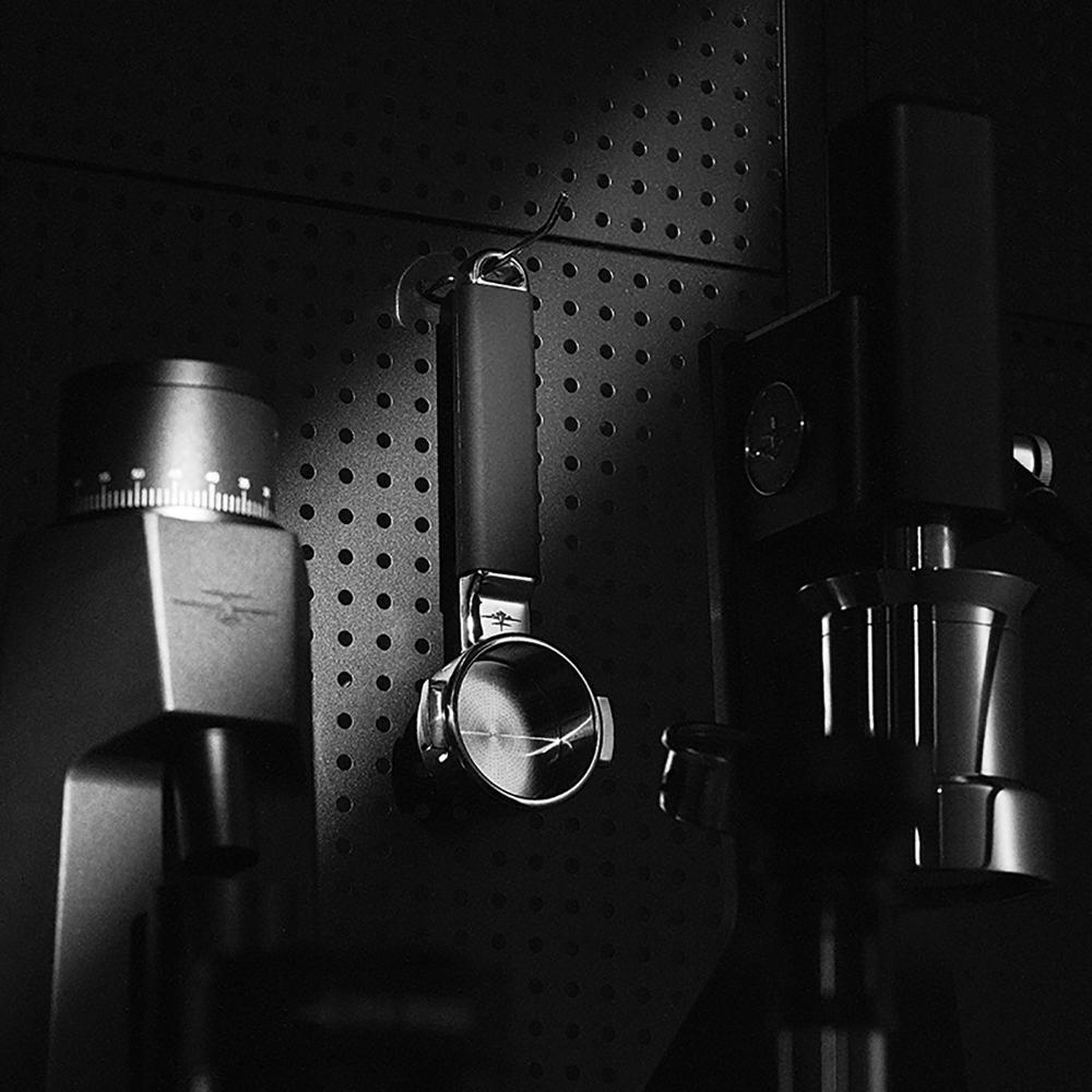 Black and white photo of coffee equipment on a perforated wall, featuring the MHW-3BOMBER YU Series Unibody Bottomless Portafilter 58mm and grinder, with dramatic lighting to highlight contrast and extraction efficiency.
