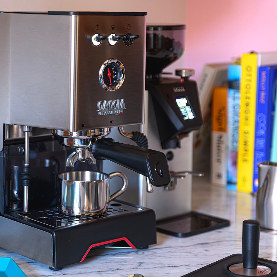 Gaggia Classic UP Semi-Automatic Espresso Machine EG2500/00 in stainless steel, featuring a 58 mm portafilter and a small metal cup on the drip tray, positioned next to a coffee grinder and a row of books.