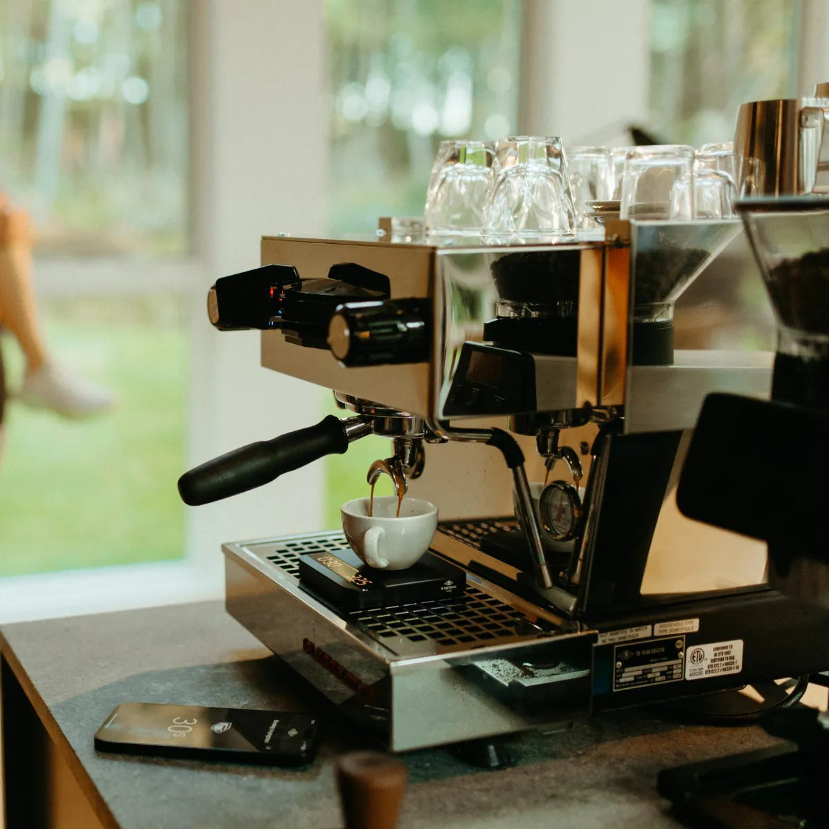 The La Marzocco Linea Mini R Dual Boiler Semi-Automatic Espresso Machine (Brushed Stainless Steel) sits on the counter, expertly brewing espresso into a white cup. Glass cups from La Marzocco are neatly arranged on top of the machine, while a phone and wristwatch—must-haves for any passionate home barista—rest nearby. Large windows in the background frame a lush green outdoor view, enhancing the scene.