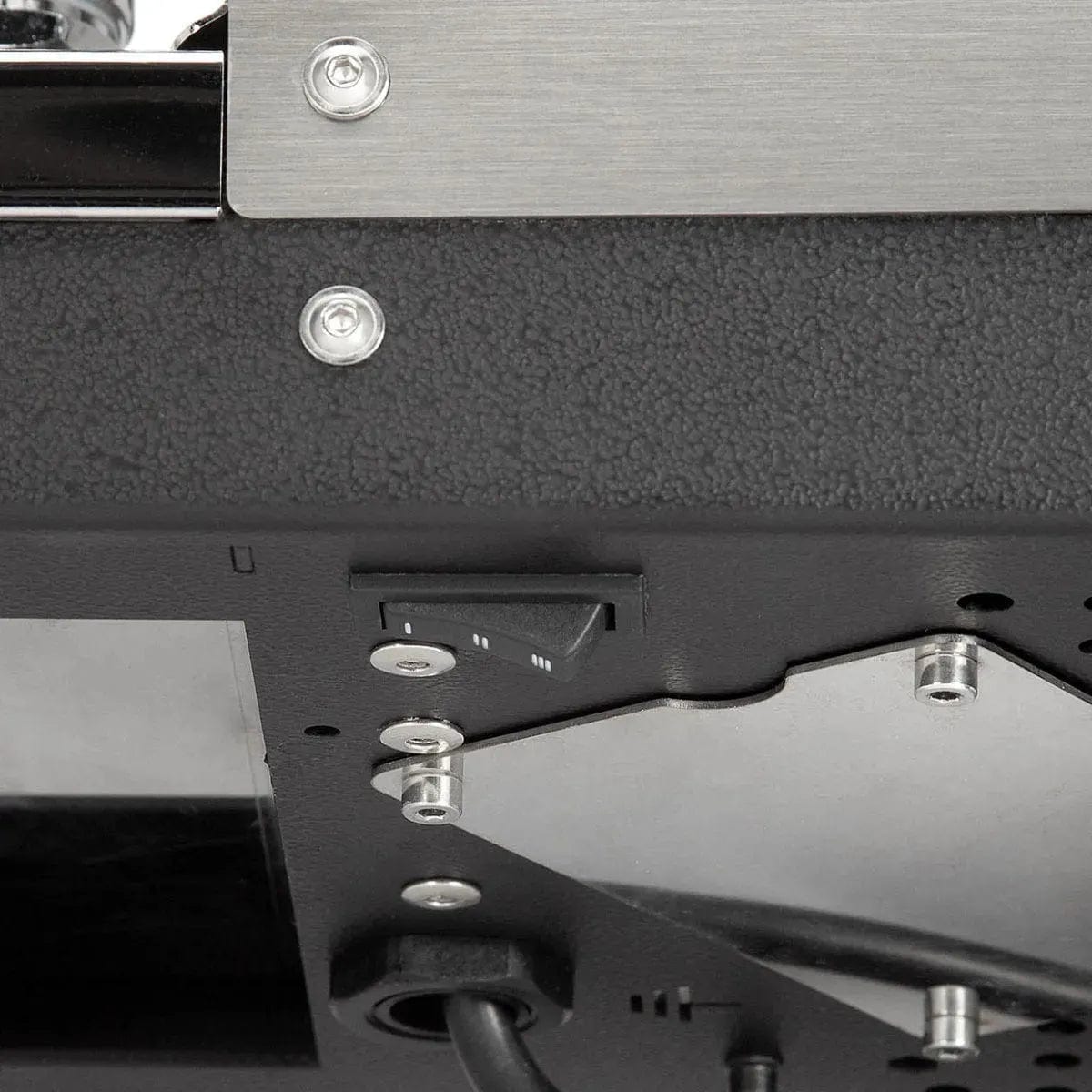 Close-up of the underside of a Profitec Jump Espresso Machine with power switch, screws, and metal panels. The black textured surface shows cables emerging below, similar to components found in a Heat Exchanger Espresso Machine.