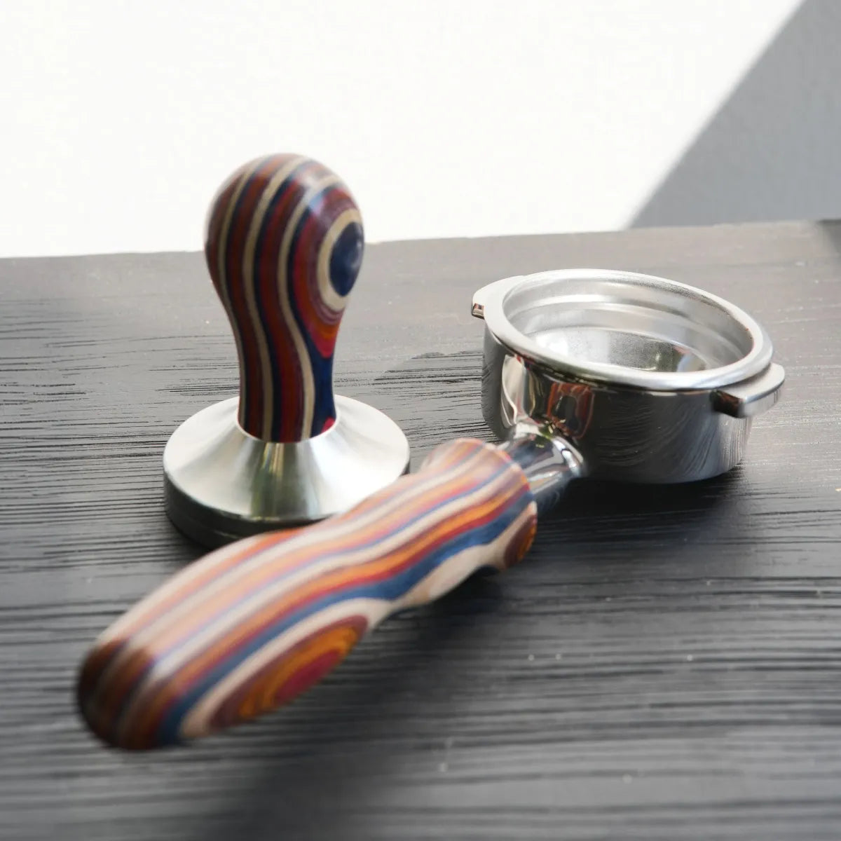 An HCS Bottomless Portafilter (Multicoloured Wood) and a matching coffee tamper for channeling detection, both featuring handles made of layered multicolored wood.