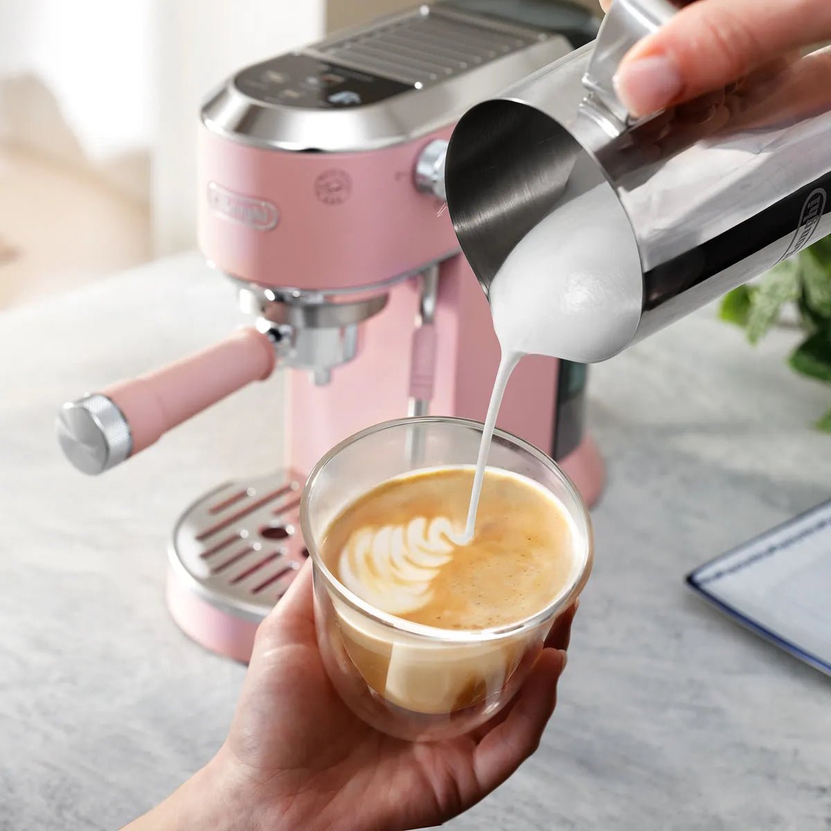 A hand pours steamed milk into a coffee glass, creating latte art using My LatteArt technology. The DeLonghi Dedica Duo Espresso Machine with Cold Brew EC890PK (Rosé) and a plant are visible in the background.