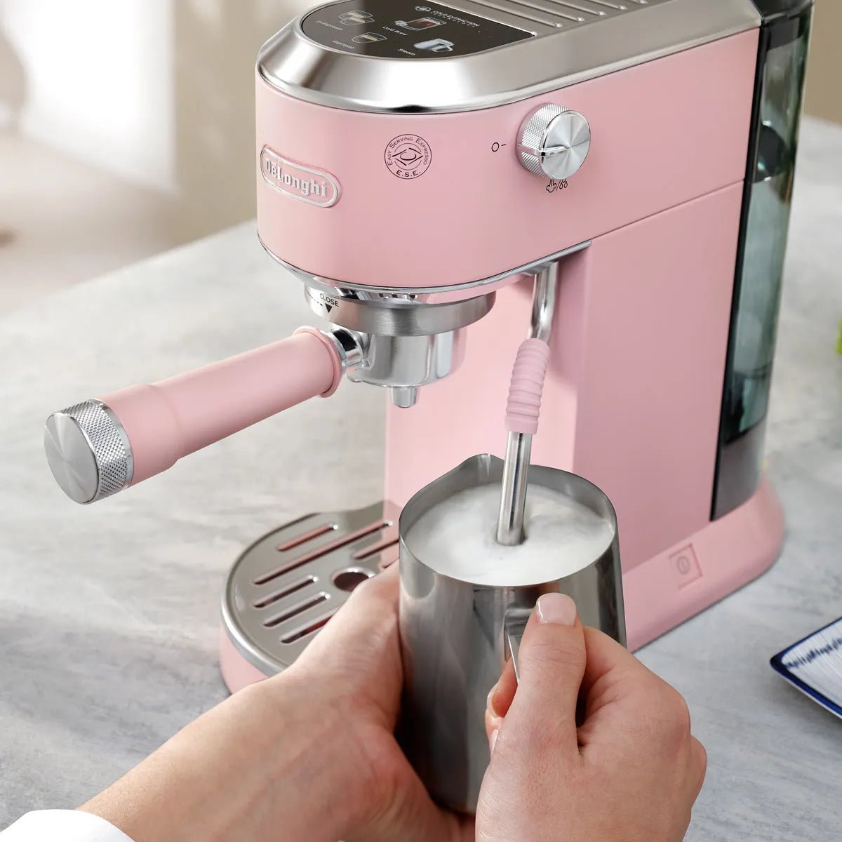 A person froths milk in a stainless steel pitcher using the DeLonghi Dedica Duo Espresso Machine with Cold Brew EC890PK (Rosé) on a light countertop.
