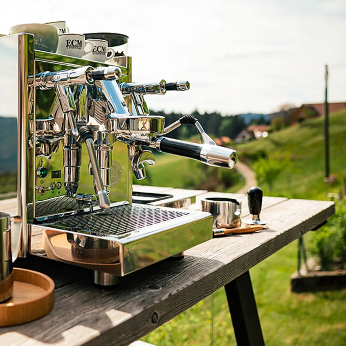 The ECM Technika VI Dual Boiler Espresso Machine with PID by ECM sits on a wooden outdoor table with coffee accessories, overlooking a lush rural landscape of hills and distant houses.