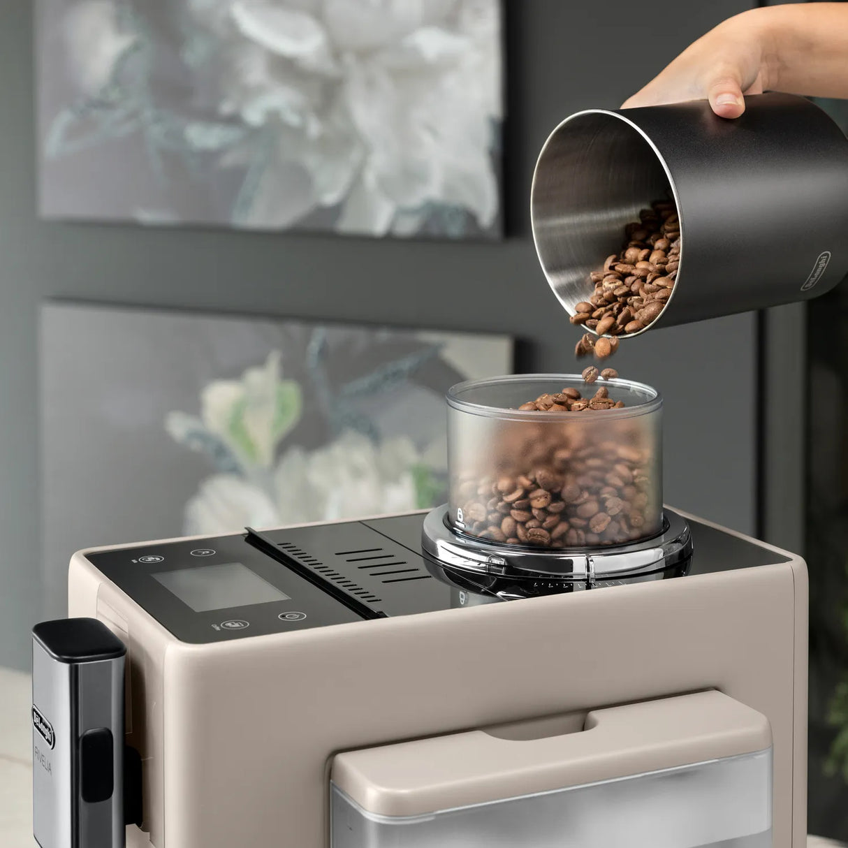 A hand pours coffee beans from a metal container into the bean hopper of the De’Longhi Rivelia Super Automatic Espresso Machine EXAM44055BG (Sand Beige) by DeLonghi, featuring the innovative Bean Switch System, with blurred white flowers in the background.