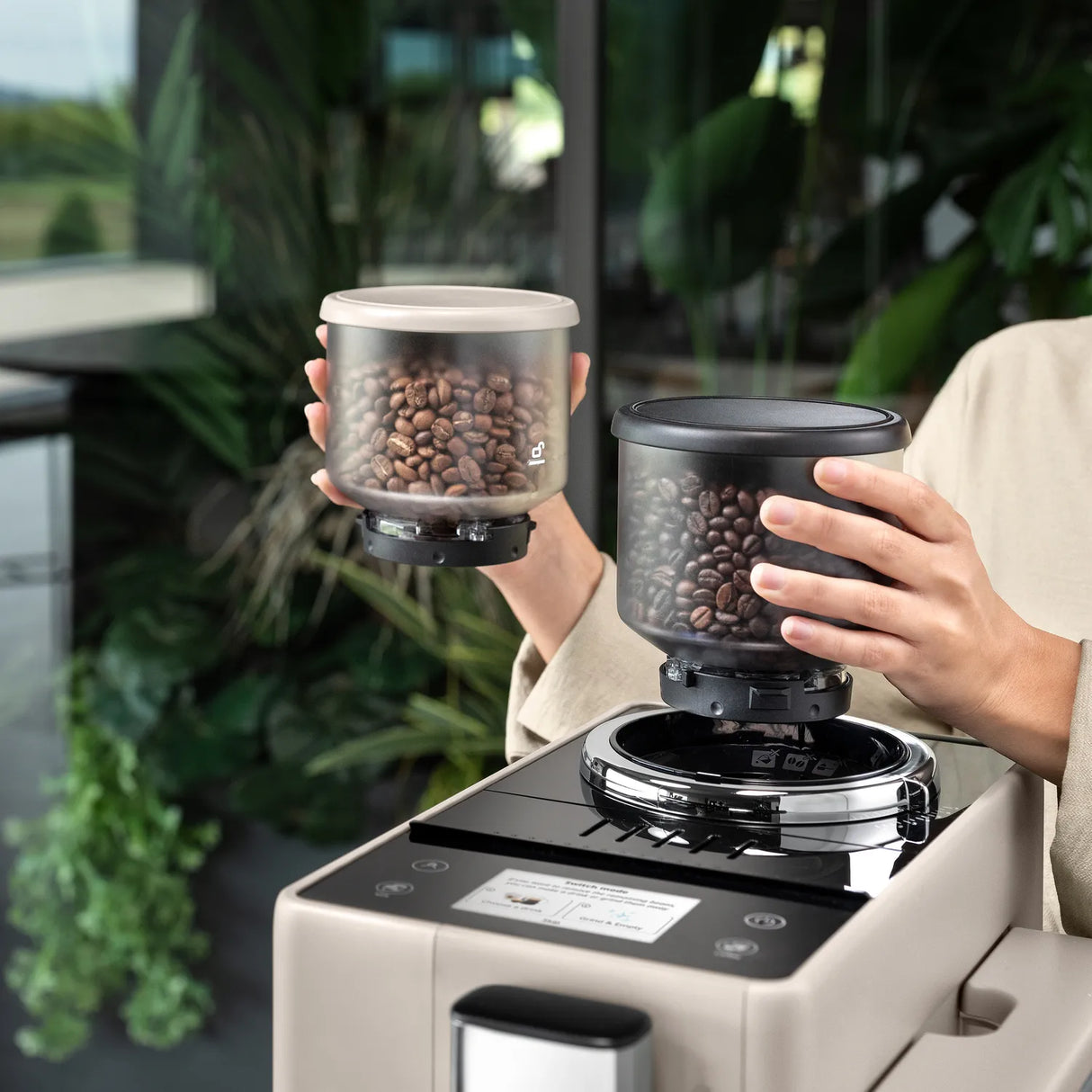 A person holds two containers filled with coffee beans, placing one onto the DeLonghi Rivelia Super Automatic Espresso Machine EXAM44055BG (Sand Beige), which features a Bean Switch System, in a bright, plant-filled room.