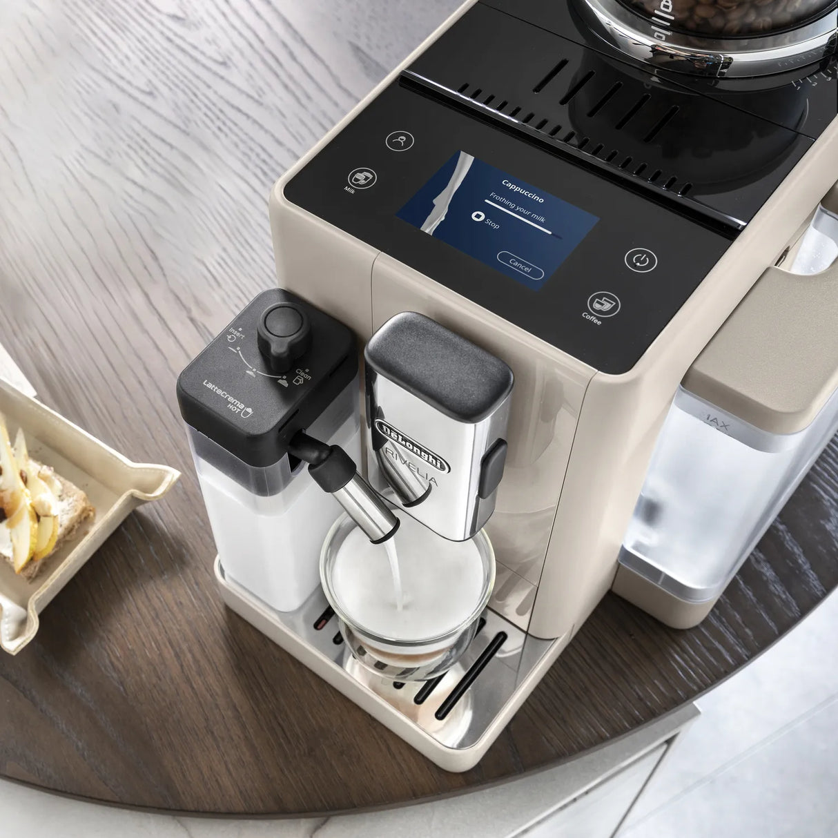A close-up of the De’Longhi Rivelia Super Automatic Espresso Machine EXAM44055BG (Sand Beige) by DeLonghi shows it frothing milk into a metal cup using LatteCrema Hot Technology to create smooth, creamy foam. The touchscreen display features coffee options, while part of a nearby table with a plate of food is visible in the frame.