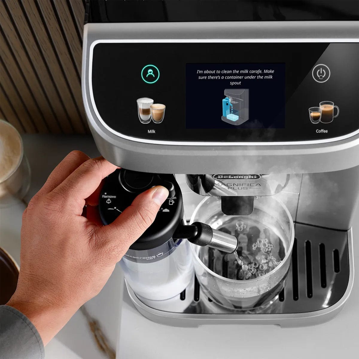 A hand adjusts the milk frother on the DeLonghi Magnifica Plus Super Automatic Espresso Machine with LatteCrema System ECAM32070SB, as cleaning instructions for the milk carafe appear on the display and coffee pours into a clear cup.