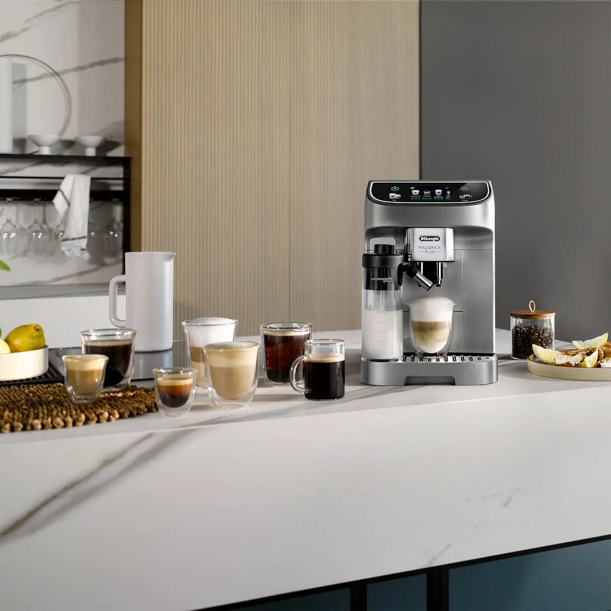 The DeLonghi Magnifica Plus Super Automatic Espresso Machine with LatteCrema System ECAM32070SB sits on a kitchen counter, surrounded by coffee drinks in clear mugs, a white pitcher, fruit, and pastries. Shelves with cups and kitchenware are visible behind it.