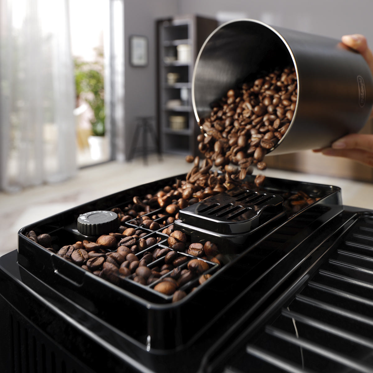 A hand pours brown coffee beans from a metal container into the bean hopper of the De'Longhi Magnifica Start Super Automatic Espresso Machine with LatteCrema System ECAM22080SB by DeLonghi, set in a bright, modern kitchen.