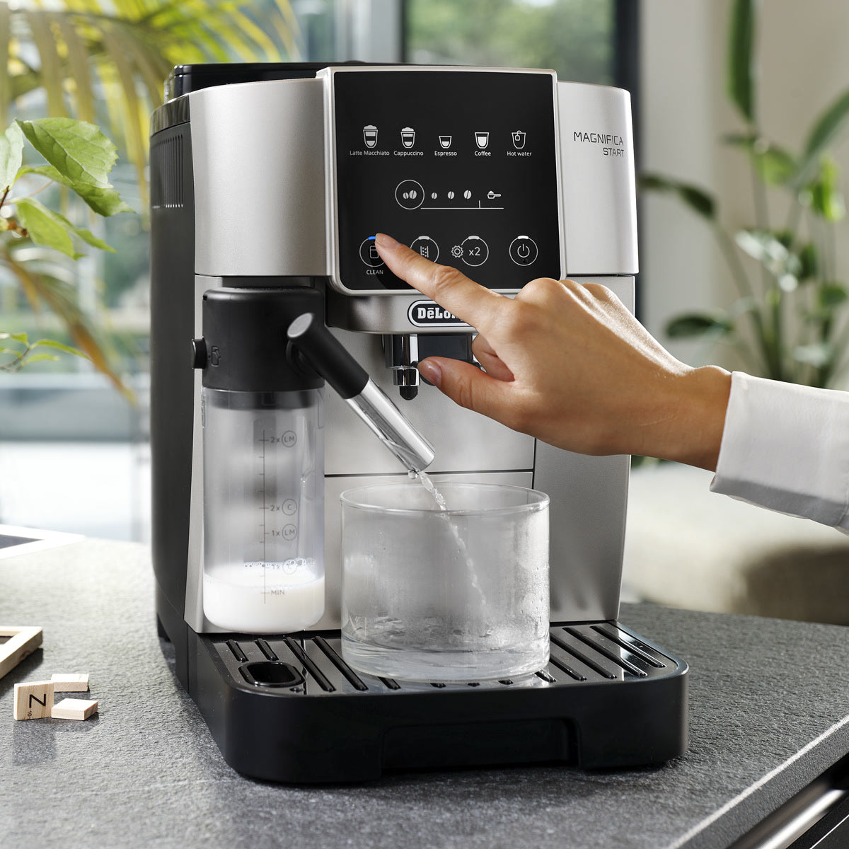A person presses a button on the De'Longhi Magnifica Start Super Automatic Espresso Machine with LatteCrema System ECAM22080SB (Silver) by DeLonghi, which has a glass under the spout and a milk container attached, sitting on a kitchen counter with plants in the background.