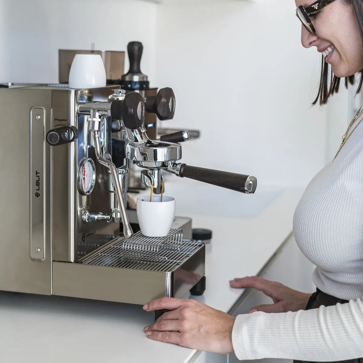 A woman in glasses and a light top smiles as she makes coffee with the Lelit Mara X3 Semi-Automatic Heat-Exchange Espresso Machine PL62X3 (Stainless Steel), watching espresso pour into a white cup on the drip tray of her modern kitchen.