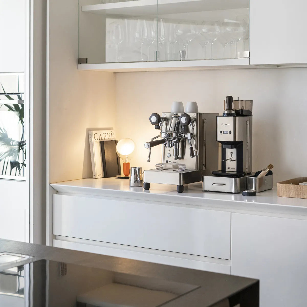 A modern kitchen counter features the Lelit Mara X3 Semi-Automatic Heat-Exchange Espresso Machine PL62X3 (Stainless Steel), a coffee grinder, small lamp, accessories, and books—all set against white cabinetry with glass shelves of glassware above.