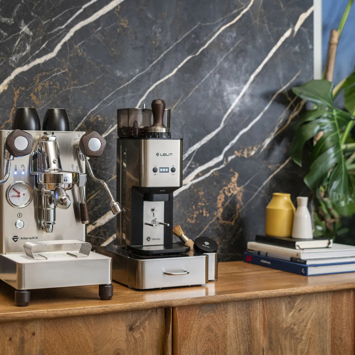 A Lelit Mara X3 Semi-Automatic Heat-Exchange Espresso Machine PL62X3 (Stainless Steel) by Lelit and a coffee grinder rest on a wooden counter before a black marble wall, alongside small vases, a plant, and stacked books.
