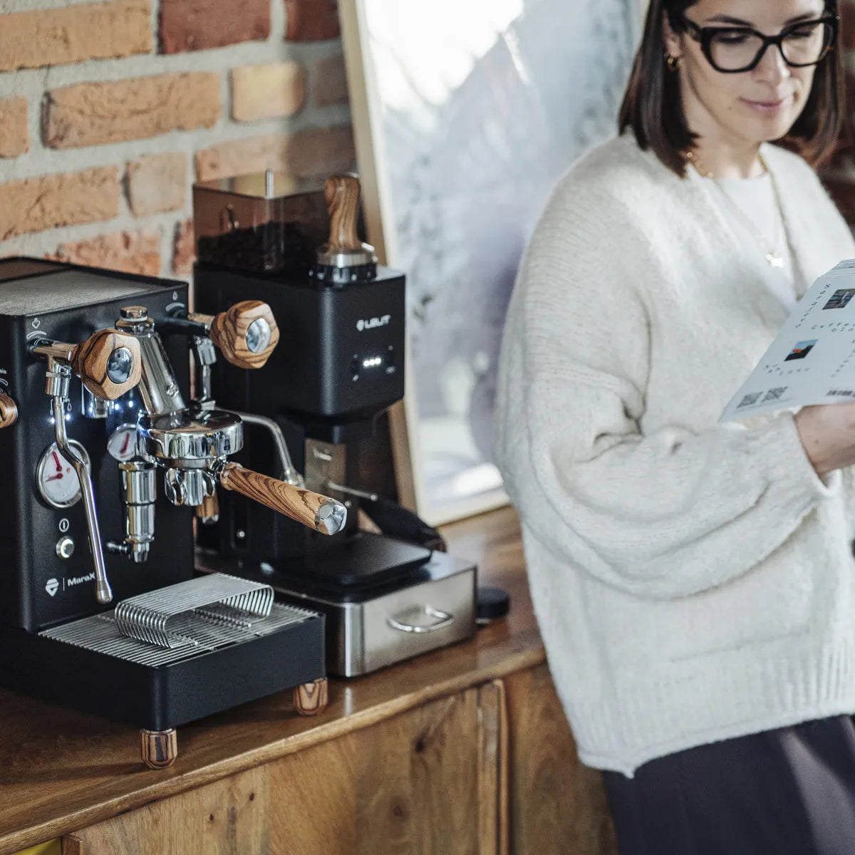 A woman in glasses and a cream sweater reads a magazine beside a black Lelit Mara X3 Semi-Automatic Heat-Exchange Espresso Machine PL62X3 and a coffee grinder on a wooden counter against a brick wall.