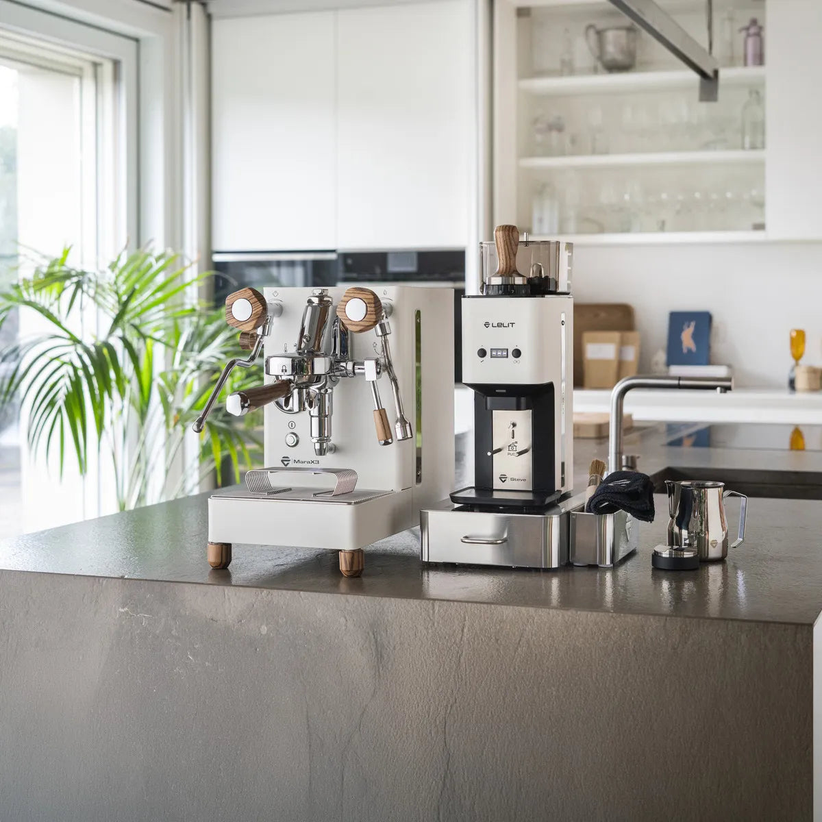 In a modern kitchen, sunlight illuminates a white Lelit Mara X3 Semi-Automatic Heat-Exchange Espresso Machine PL62X3 by Lelit, along with a coffee grinder, milk frother, and kettle on a gray countertop beside a green plant.