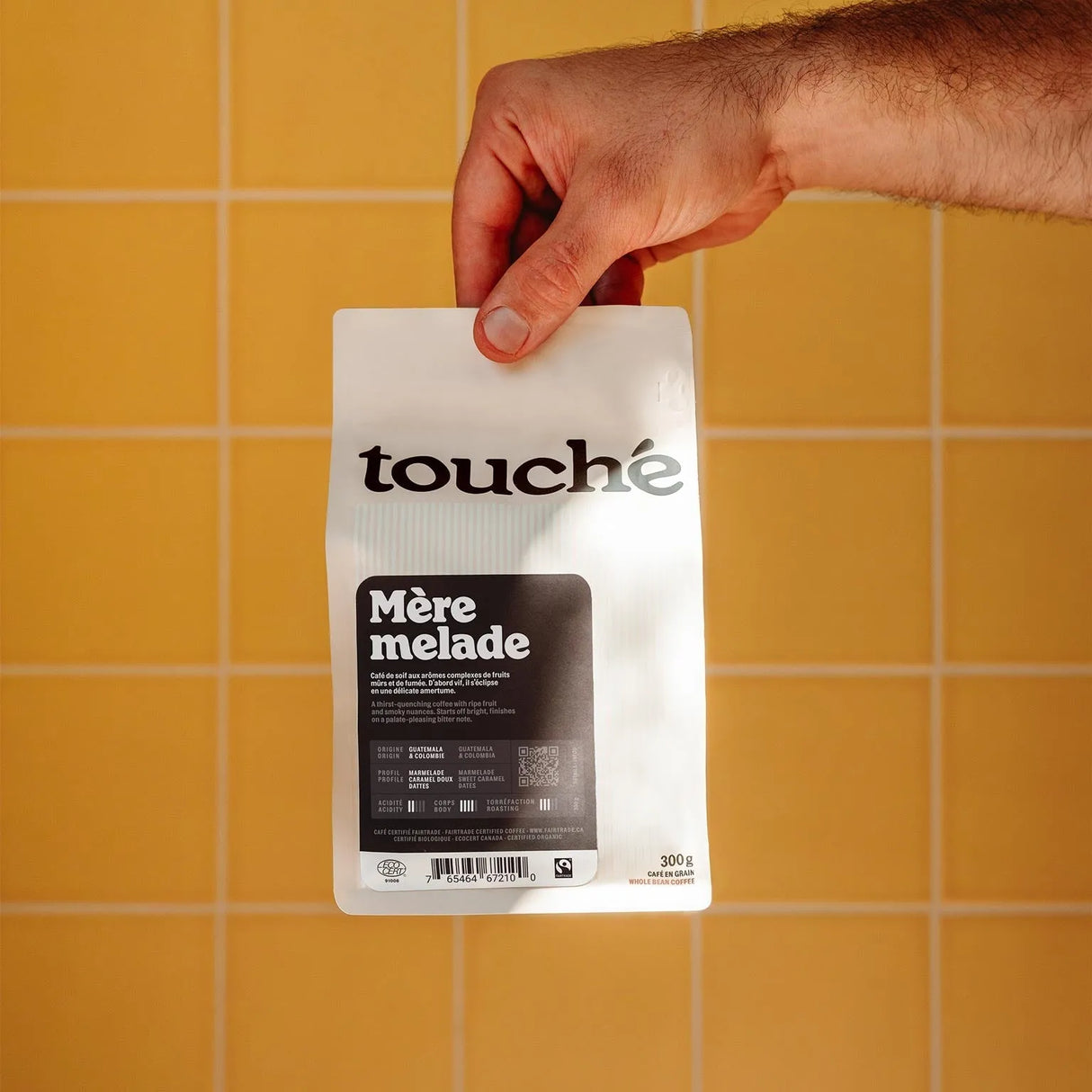 A hand holds a bag of Cafe Touche Mere Melade Whole Bean Coffee (1 KG) by Cafe Touche, featuring organic medium roast beans, set against a yellow tiled background.