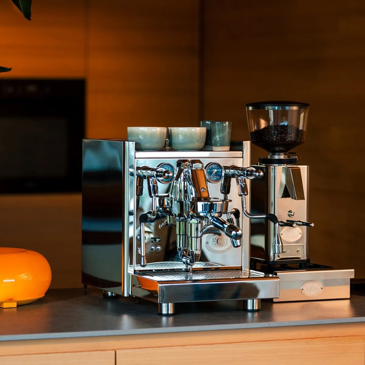 The ECM Technika VI Dual Boiler Espresso Machine w/ PID by ECM, topped with two cups, sits on a kitchen counter beside a coffee grinder filled with beans, set against warm wooden cabinetry.