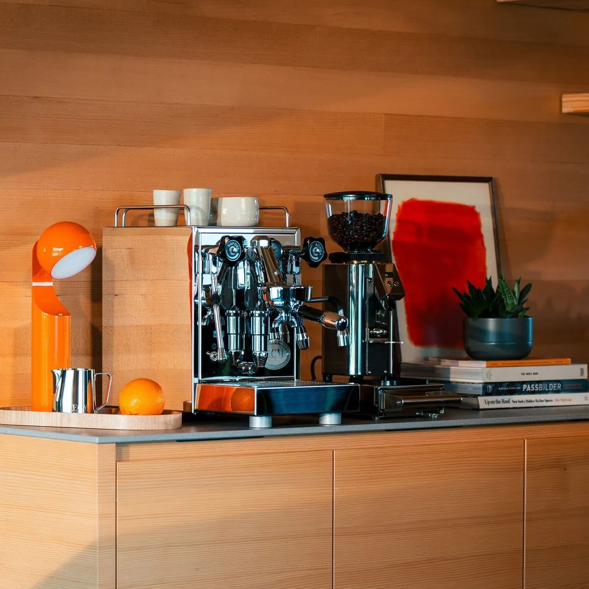 A modern coffee station on a wooden counter showcases the ECM Technika VI Dual Boiler Espresso Machine with PID by ECM, a coffee grinder, orange lamp, cups, oranges, books, framed abstract art, and a small potted plant.