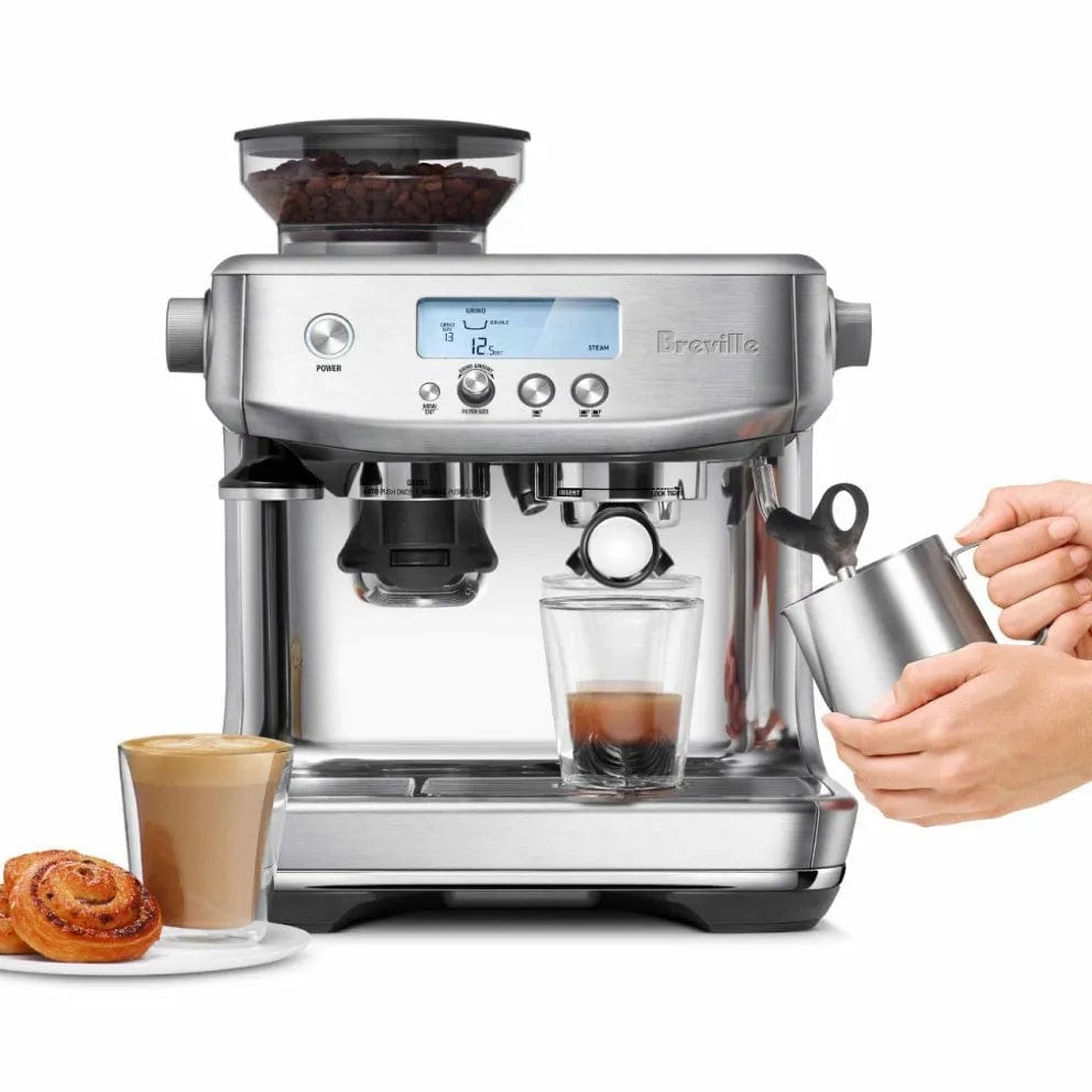 A black stainless steel Breville The Barista Pro Espresso Machine BES878BST with a digital display and built-in coffee grinder is making espresso. A hand is frothing milk in a metal jug. A glass of layered latte and a cinnamon roll are placed next to the semi-automatic espresso machine with its efficient ThermoJet heating system.