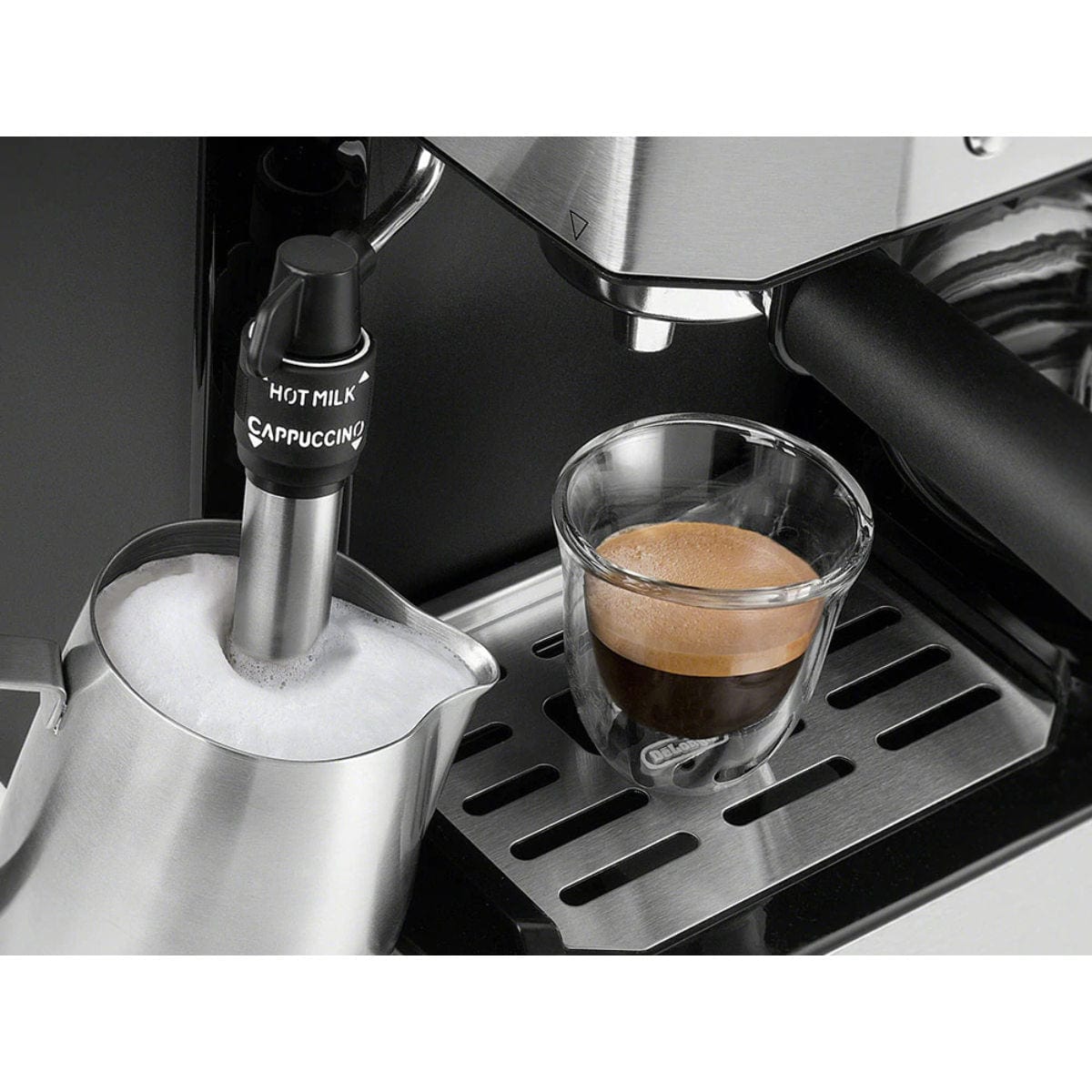 A close-up of the DeLonghi Combination All-in-One Coffee, Espresso & Latte Machine COM532M shows it brewing espresso into a glass cup on the drip tray, while the milk frother steams milk in a metal pitcher beside it.