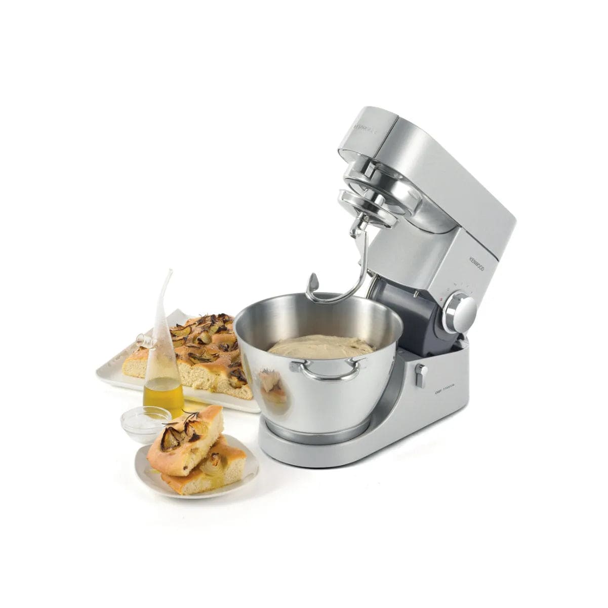 A Kenwood Chef Titanium KMC011 Stand Mixer & Kitchen Machine with a 720 Watt motor and a spacious 4.7L stainless steel bowl sits on a white surface, equipped with expandable attachments. Beside it, a plate of freshly baked bread topped with mushrooms and a glass bottle of olive oil complete the culinary scene.