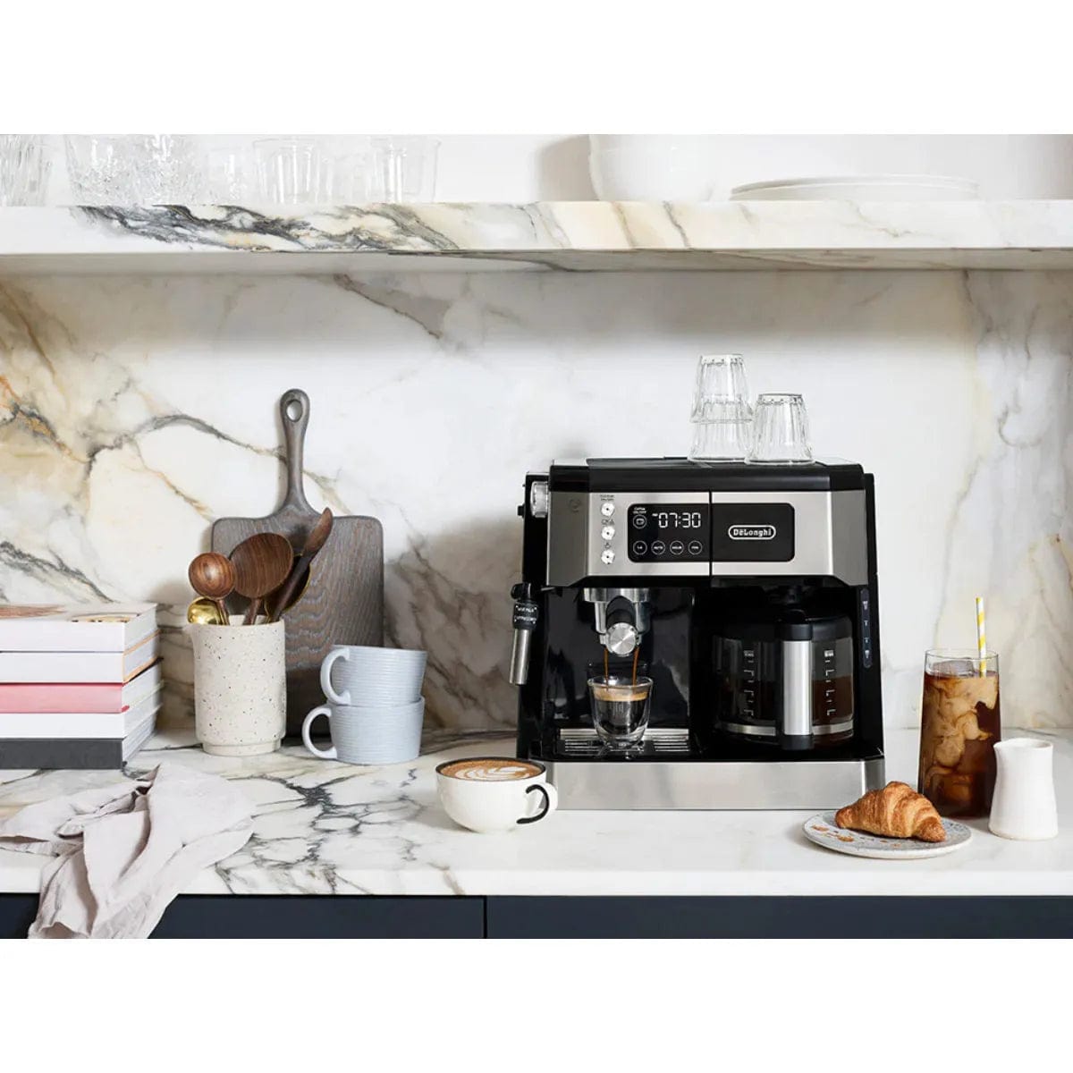 A DeLonghi Combination All-in-One Coffee, Espresso & Latte Machine COM532M sits on a marble countertop, surrounded by mugs, an iced coffee, a foamy latte, a croissant on a plate, glassware, stacked books, and kitchen utensils.