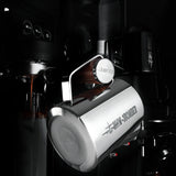 A MHW-3BOMBER Latte Art Trio Bundle pitcher labeled “49MM BOMBER” is attached to an espresso machine, pouring coffee into a cup in a sleek, modern setting.