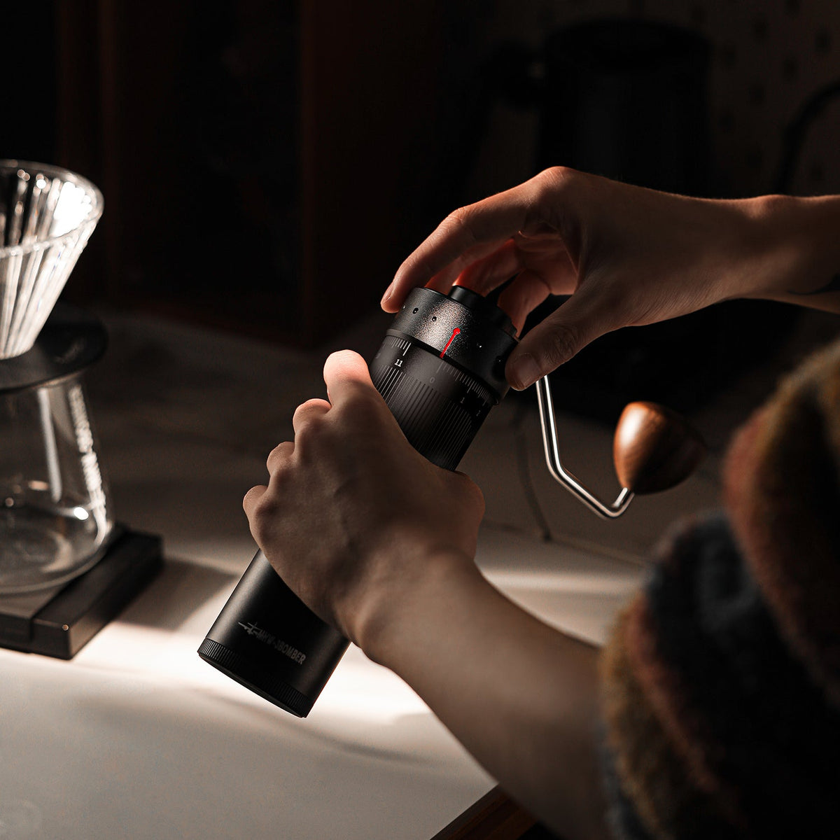 A person adjusts the MHW-3BOMBER Precision R3 Pro Manual Coffee Grinder (Black) with both hands on a kitchen counter beside a coffee dripper, warm light highlighting their hands and the grinder.