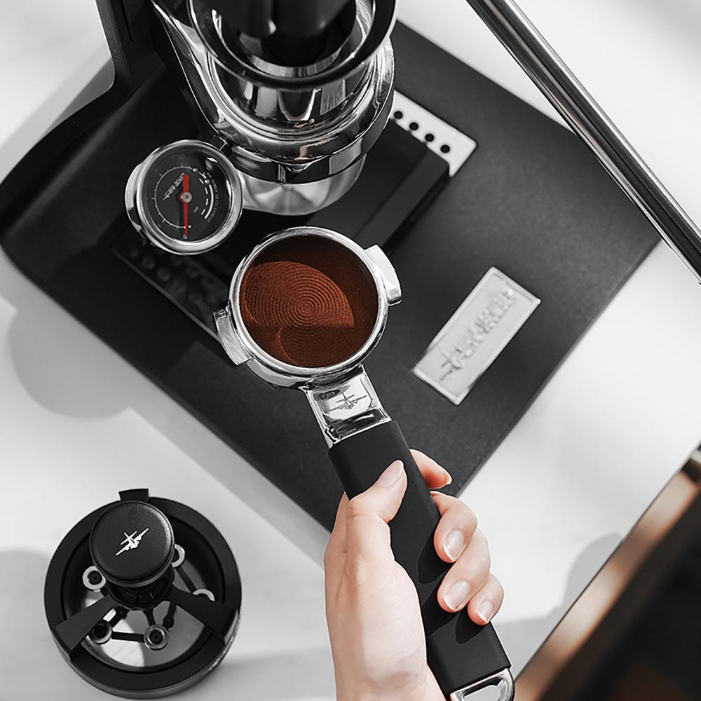 A hand holds the MHW-3BOMBER YU Series Unibody Bottomless Portafilter 58mm with ground coffee under a manual espresso machine, alongside a pressure gauge and tamper on a black base for optimal extraction.