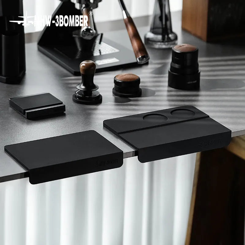 A sleek coffee bar setup with black barista tools, featuring the MHW-3BOMBER Silicone Corner Tamping Mat SP5312B (Black) on a gray counter alongside tampers, a scale, espresso tools, and a portafilter.