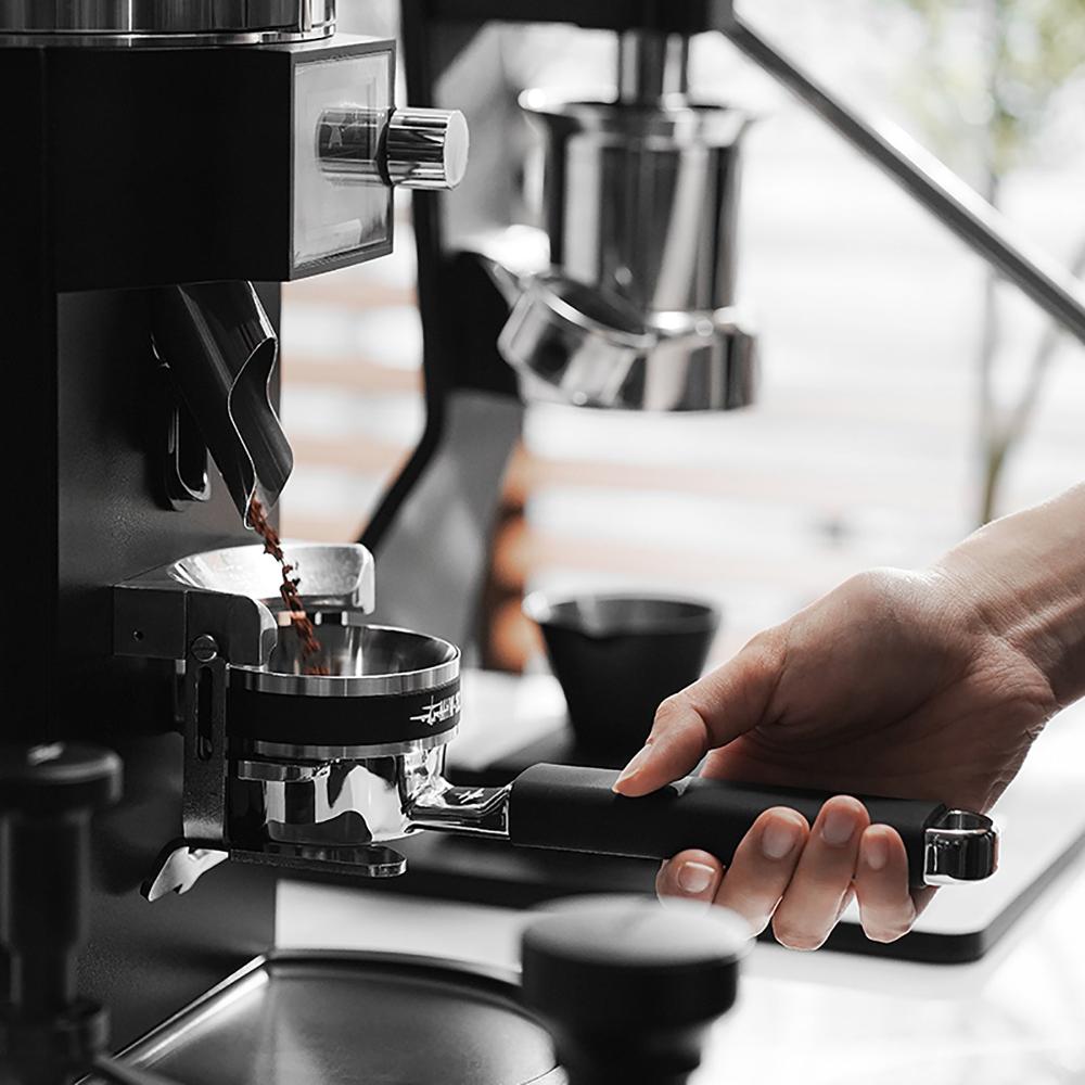 A hand holds the MHW-3BOMBER YU Series Unibody Bottomless Portafilter 58mm under an espresso machine as coffee pours through precise filter holes, emphasizing extraction efficiency against a blurred backdrop.