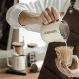 A barista in a brown apron pours steamed milk from a MHW-3BOMBER Latte Art Milk Pitcher 5.0 500ml (White) into coffee, creating delicate latte art. Coffee equipment and another drink are blurred in the background.