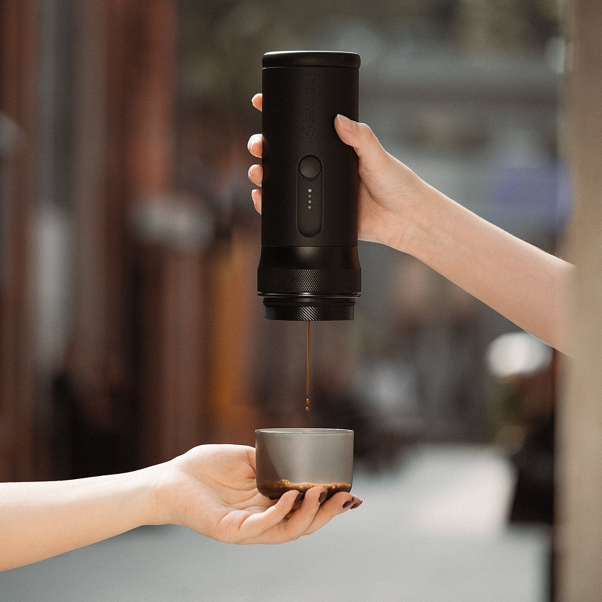 Two hands: one operates the MHW-3BOMBER X-press58 Portable Espresso Machine CM6954 (Black) pouring coffee into a cup, with a blurred outdoor adventure scene in the background.
