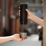 Two hands: one operates the MHW-3BOMBER X-press58 Portable Espresso Machine CM6954 (Black) pouring coffee into a cup, with a blurred outdoor adventure scene in the background.