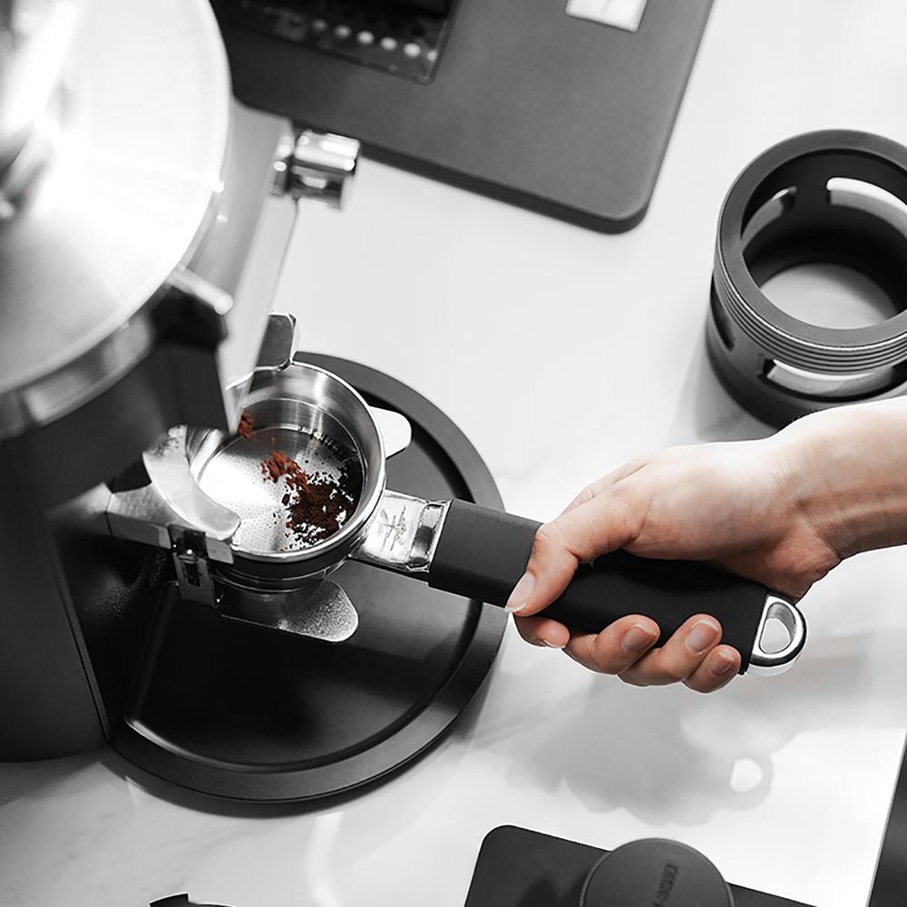 A hand holds the MHW-3BOMBER YU Series Unibody Bottomless Portafilter 58mm, filled with ground coffee, placing it into an espresso machine on a countertop surrounded by coffee accessories.
