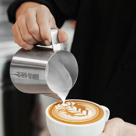 Someone pours steamed milk from the MHW-3BOMBER Latte Art Milk Pitcher 5.0 500ml (Stainless Steel) into coffee, crafting a leaf pattern on top.